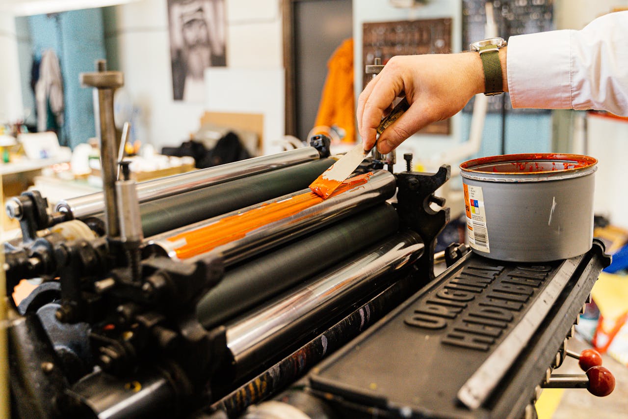 gallery-3 A detailed view of a hand applying orange ink to a vintage printing press roller indoors.
