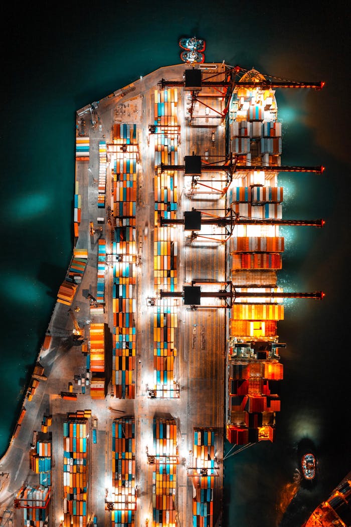 gallery-6 Dramatic aerial view of a busy shipping port with colorful containers and bright lights at night.